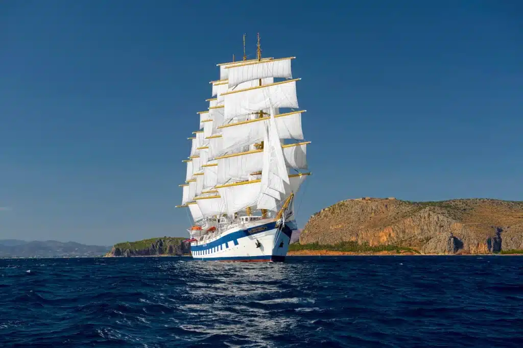 Het indrukwekkende vijfmaster zeilschip Royal Clipper van Star Clippers met gehesen witte zeilen, varend op een diepblauwe zee langs een bergachtige kustlijn.