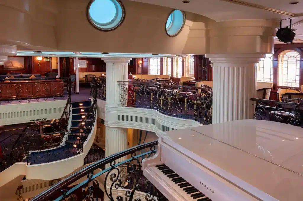 De sfeervolle Piano Bar aan boord van de Royal Clipper, met een witte vleugel op de voorgrond en zicht op de klassieke trap en galerij met patrijspoorten.