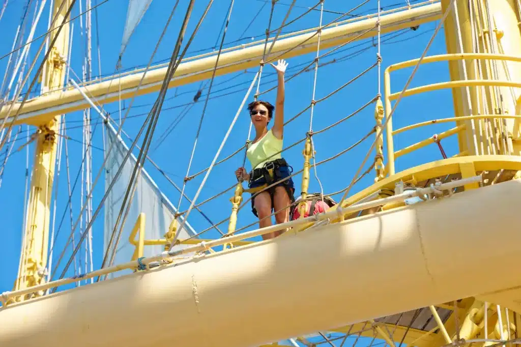 Een enthousiaste gast in een veiligheidsharnas zwaait vanaf de ra van de Royal Clipper, met de indrukwekkende masten en tuigage tegen een strakblauwe lucht.
