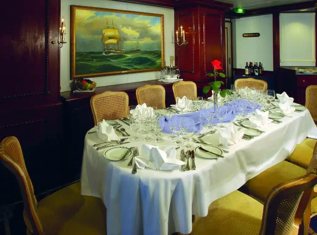 Een elegant gedekte tafel in de dining room van de Star Clipper, met wit linnen, blauwe accenten, kristallen glazen en een klassiek maritiem schilderij aan de houten wand.