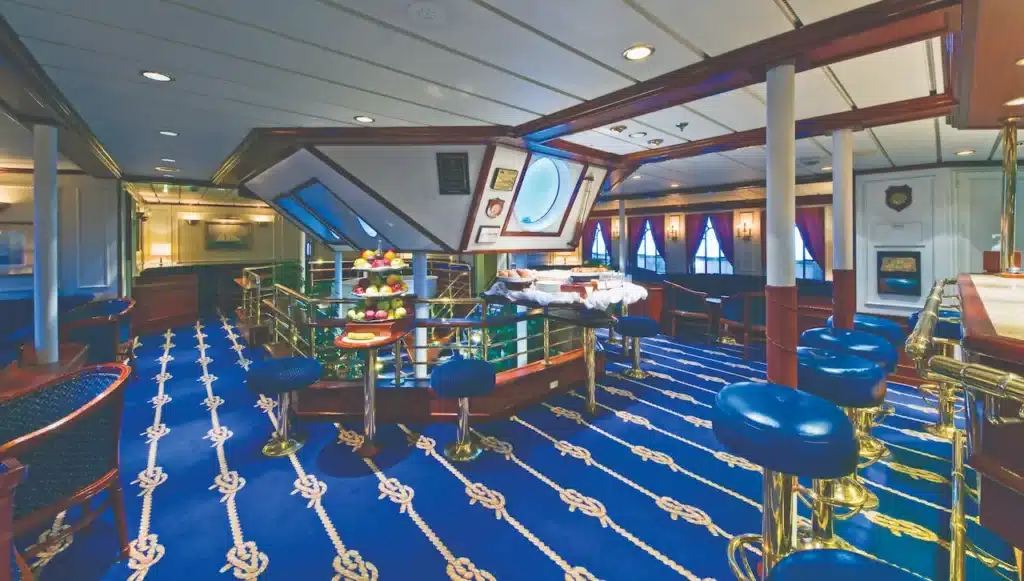 Het elegante interieur van de Piano Bar en Salon op de Star Clipper, met wit gestreepte banken, een witte piano, klassieke houten pilaren en gasten die ontspannen in een lichte ambiance.