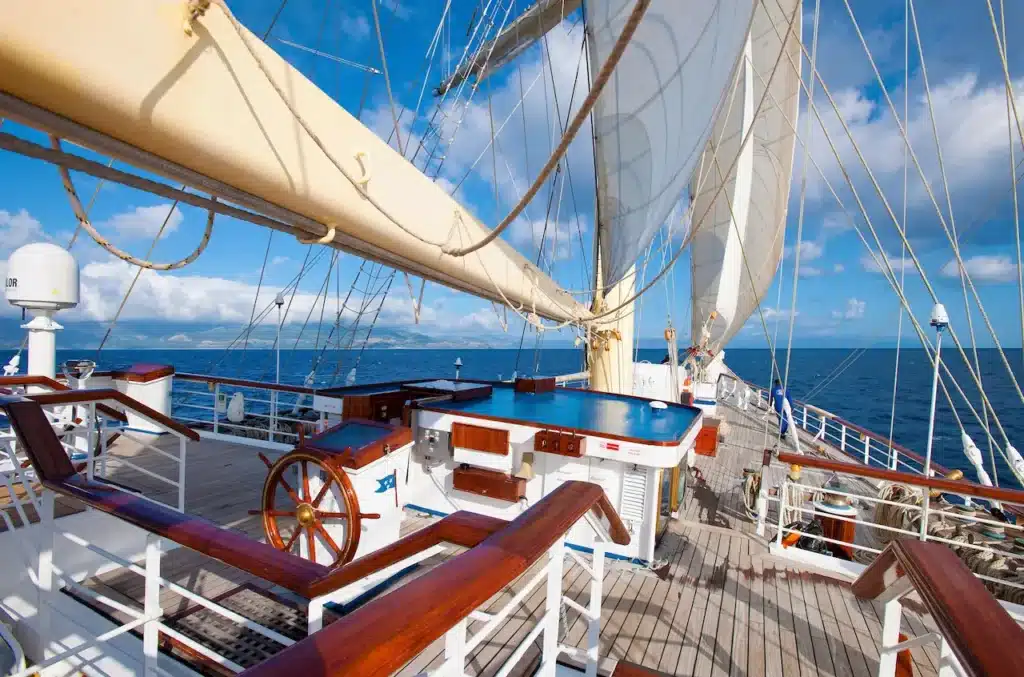 Een houten klassiek stuurwiel op het open dek van de Star Clipper, met uitzicht over de reling op de blauwe zee en de witte zeilen onder een lichtbewolkte hemel.