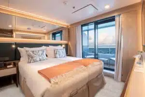 Luxe tweepersoonsbed in de Deluxe Veranda Suite op een Atlas Ocean Voyages schip met een glazen schuifdeur naar het privébalkon.