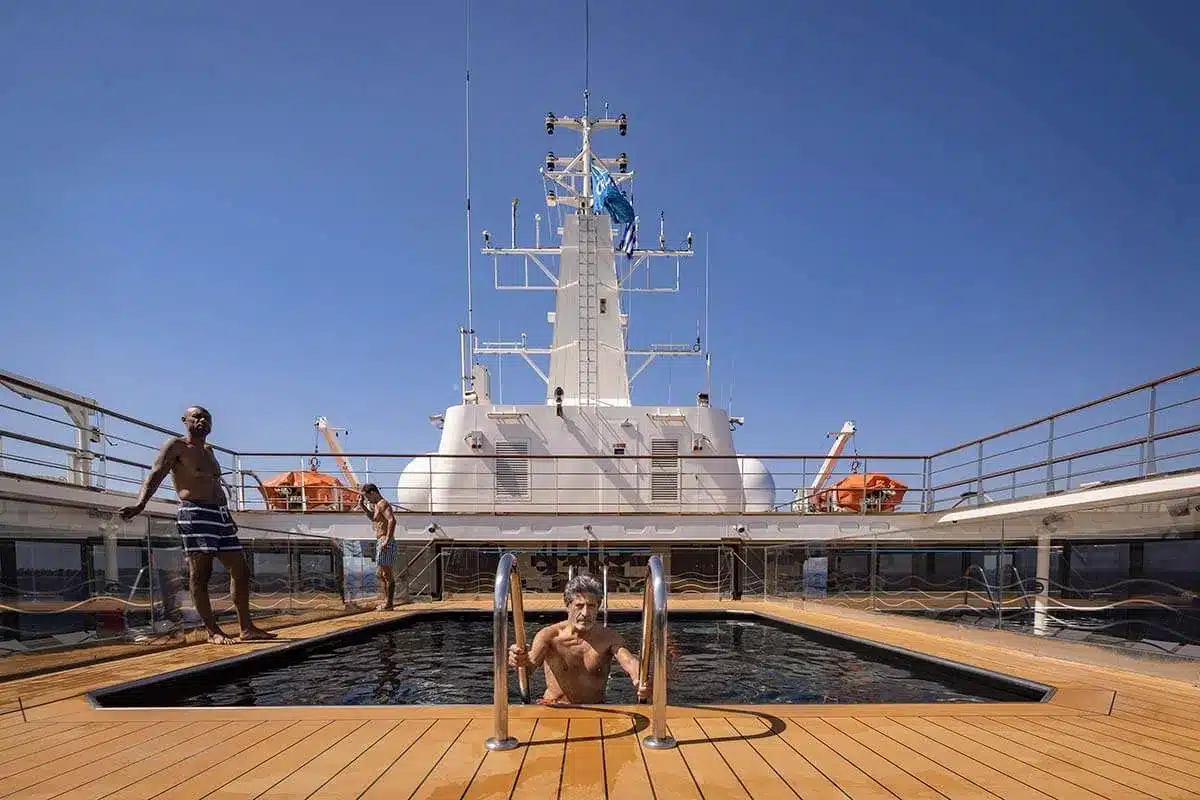 Gasten bij het zwembad op het dek van een cruiseschip onder een strakblauwe hemel.