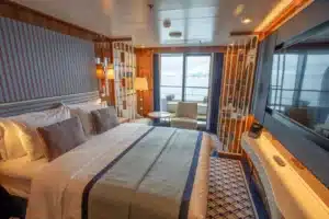 Luxe slaapkamer van de Horizon Suite (H1/H2) op de World Traveller met een gestreept hoofdbord, zithoek en uitzicht op zee door het verlaagde balkonvenster.