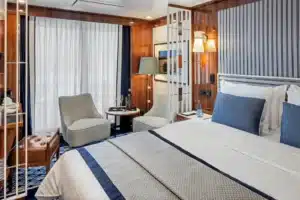 Luxe slaapkamer van de Veranda Suite (V1/V2) op de World Traveller met een gestreept hoofdbord, zithoek en gordijnen die leiden naar het privébalkon.