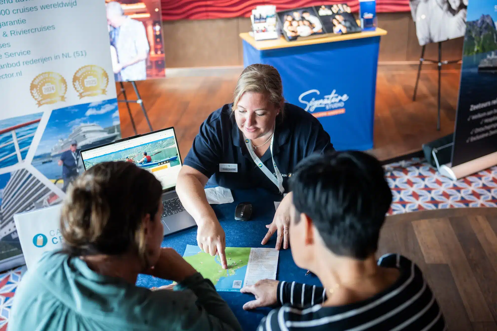 Reisbegeleider van Captain Cruise geeft persoonlijk advies aan reizigers aan tafel met routekaart en laptop tijdens een cruise evenement