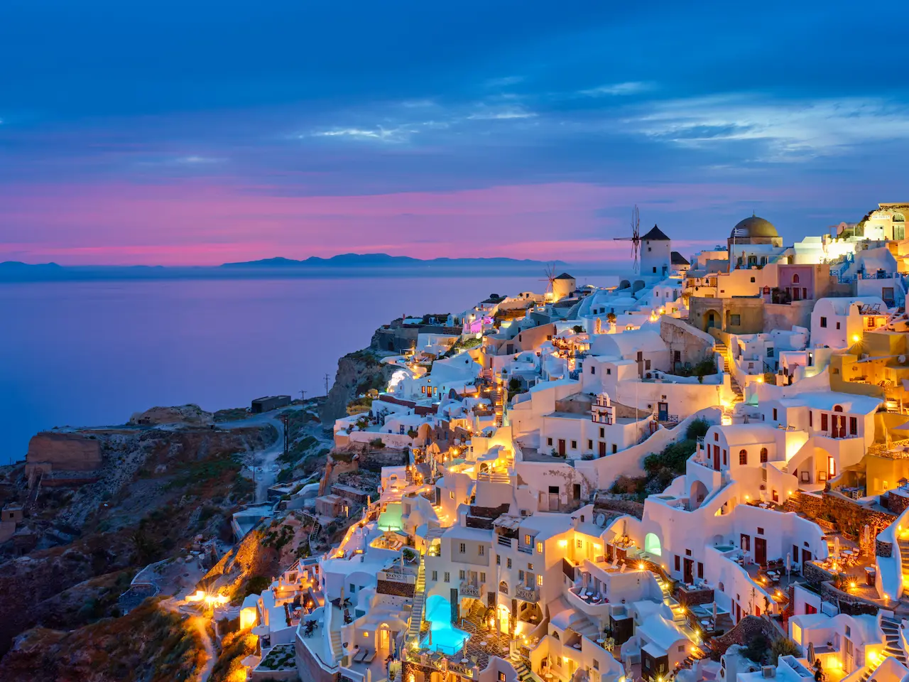 Panoramisch uitzicht op Oia, Santorini in Griekenland tijdens de avondschemering met verlichte witte huizen, blauwe koepels en een molen langs de kliffen.